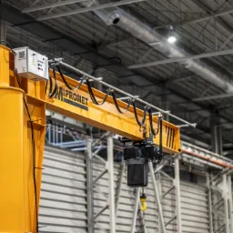 Żółty żuraw słupowy z napisem 'PROMET' w hali produkcyjnej, widoczne elementy sterowania i hak