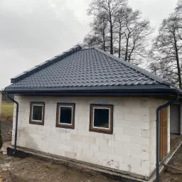 Nowy, grafitowy dach z blachodachówki modułowej na surowym budynku z bloczków, widoczne rynny i okna w trakcie montażu.