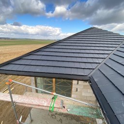 Juko System Adrian Kotomski - Fragment dachu z czarnej dachówki modułowej, widok z rusztowania na niezabudowany budynek w tle pola i pochmurnego nieba.