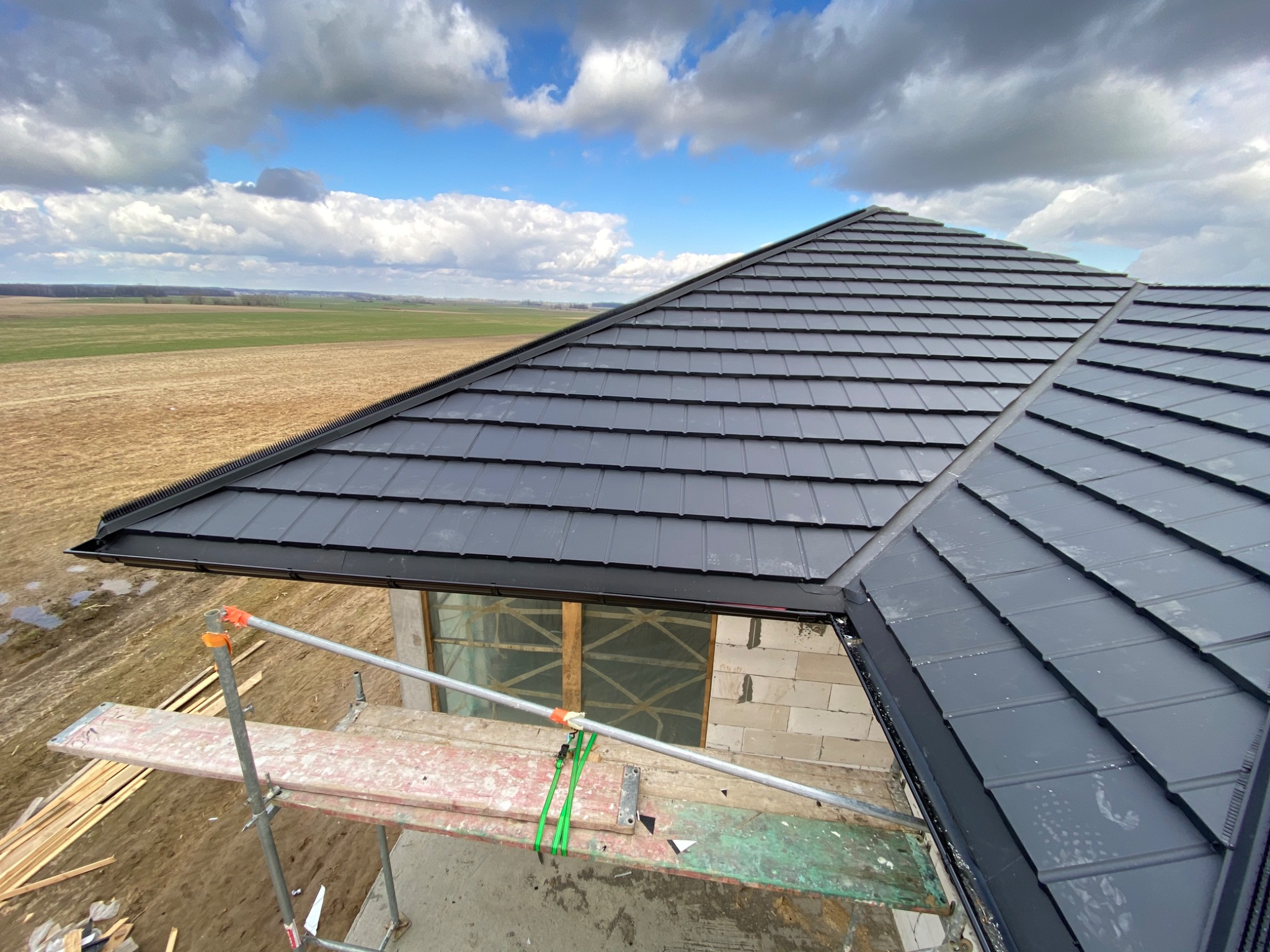 Fragment dachu z czarnej dachówki modułowej, widok z rusztowania na niezabudowany budynek w tle pola i pochmurnego nieba.