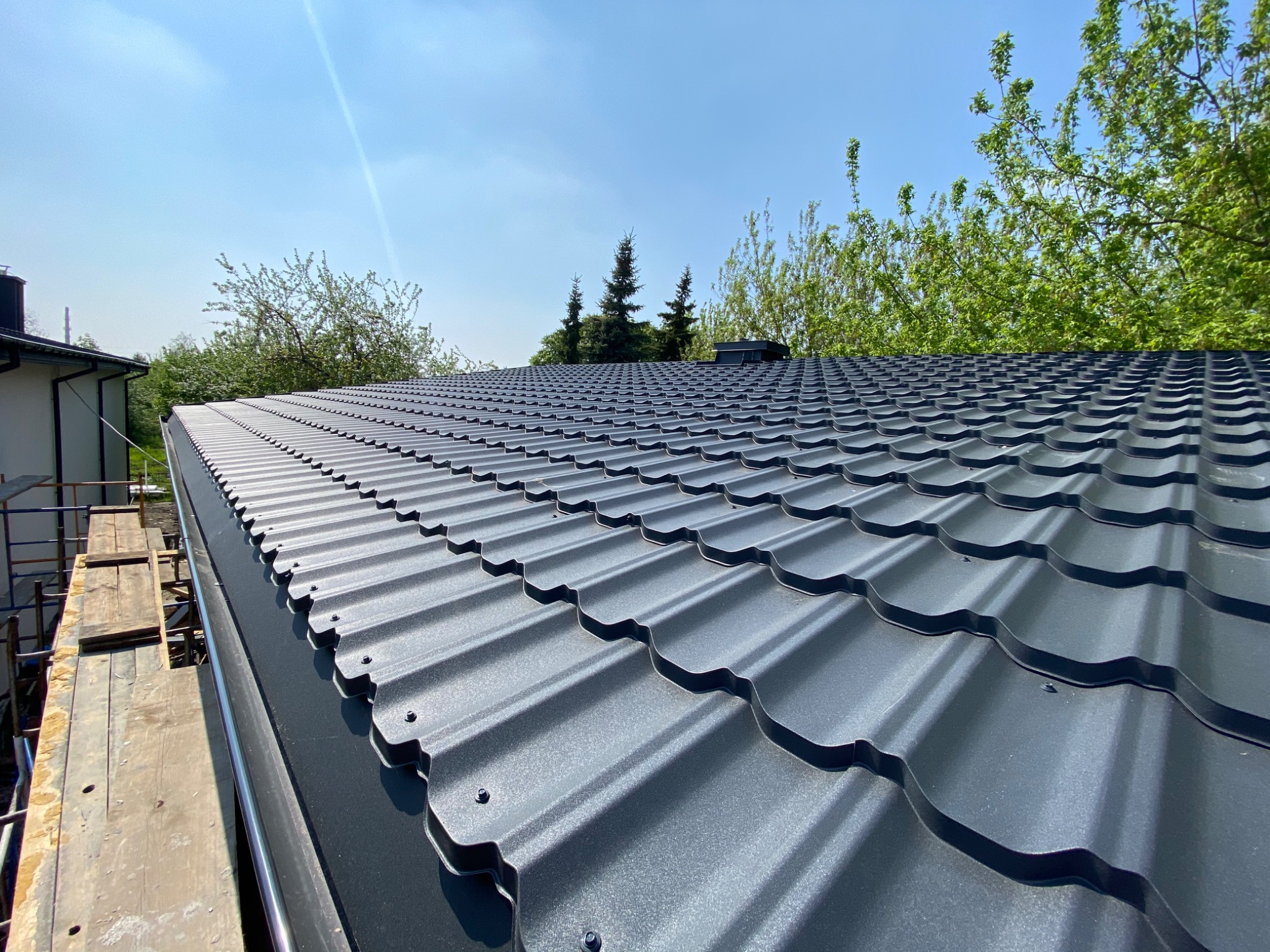 Nowy, ciemnoszary dach z blachy trapezowej widziany z dołu, z widocznym fragmentem rusztowania i zielenią drzew w tle na tle błękitnego nieba.
