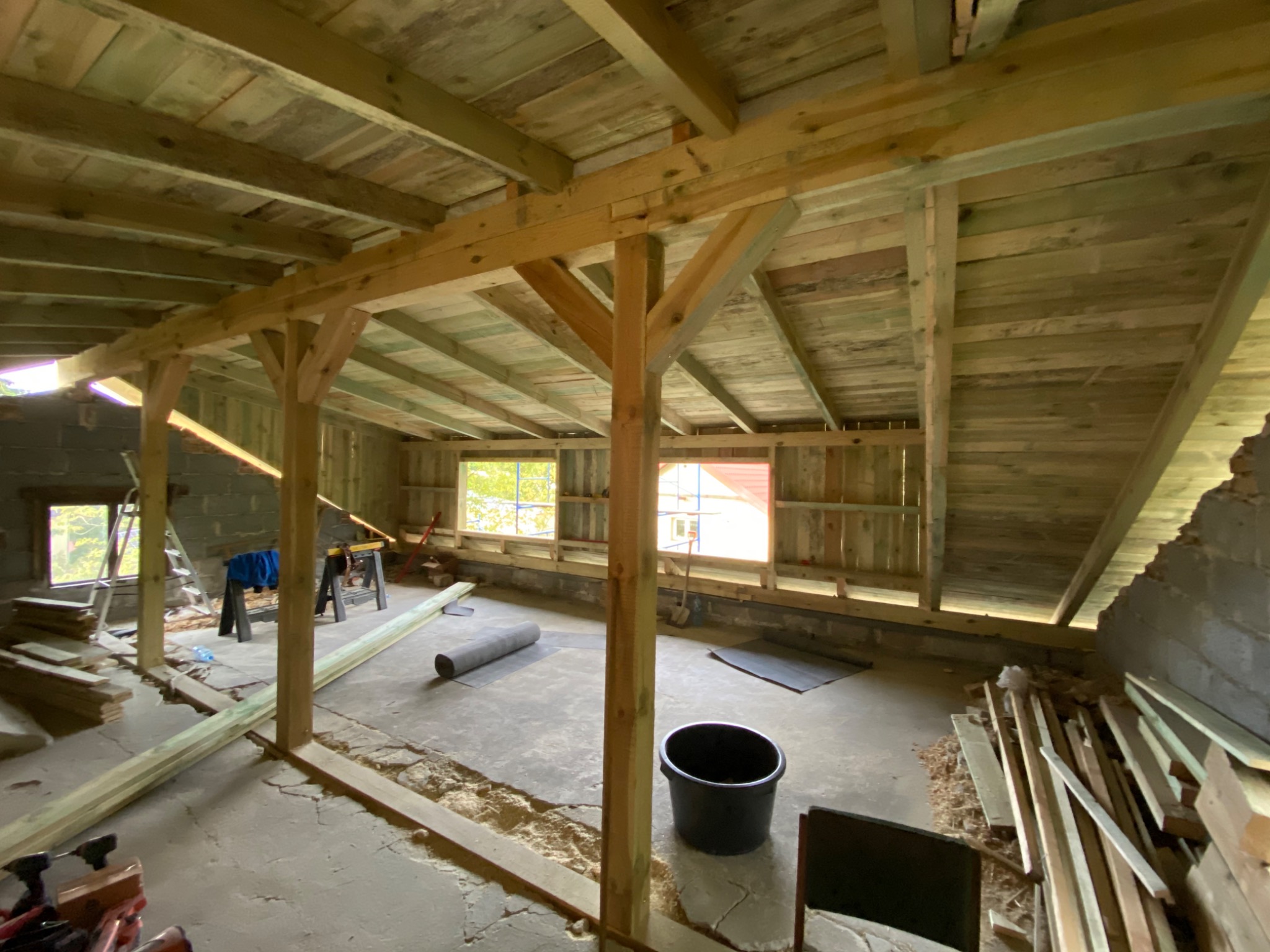 Wnętrze budynku w trakcie budowy więźby dachowej, widoczne drewniane elementy konstrukcyjne, okna, materiały budowlane i narzędzia.
