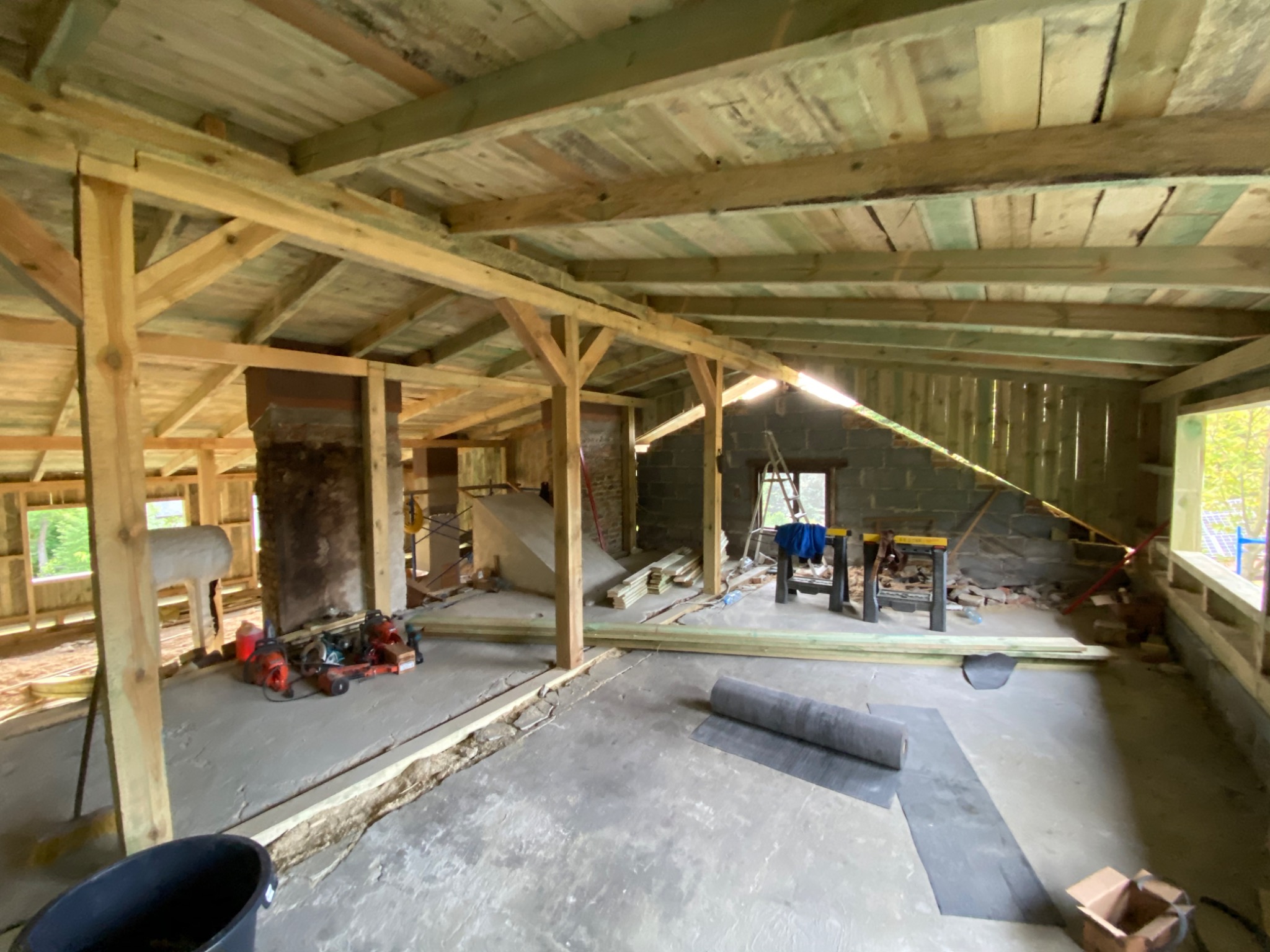 Wnętrze budynku w trakcie budowy z widoczną drewnianą konstrukcją dachu, częściowo pokrytą deskami, kominem, narzędziami i materiałami budowlanymi na betonowej podłodze.