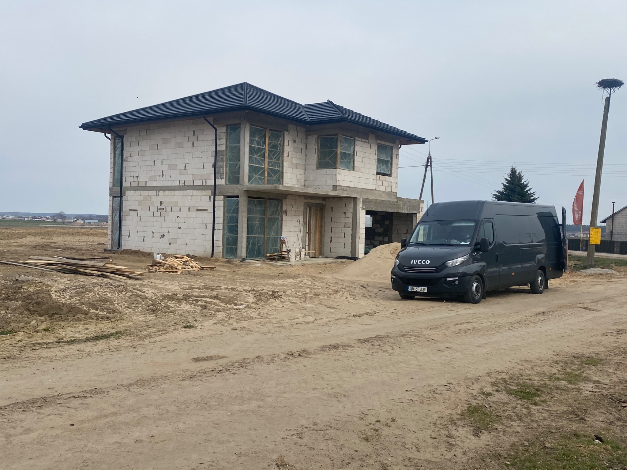 Surowy stan domu jednorodzinnego z ciemną dachówką, okna zabezpieczone folią, na posesji zaparkowany czarny bus Iveco, w tle widoczne pola i łąki, po prawej stronie słup z gniazdem bocianim.