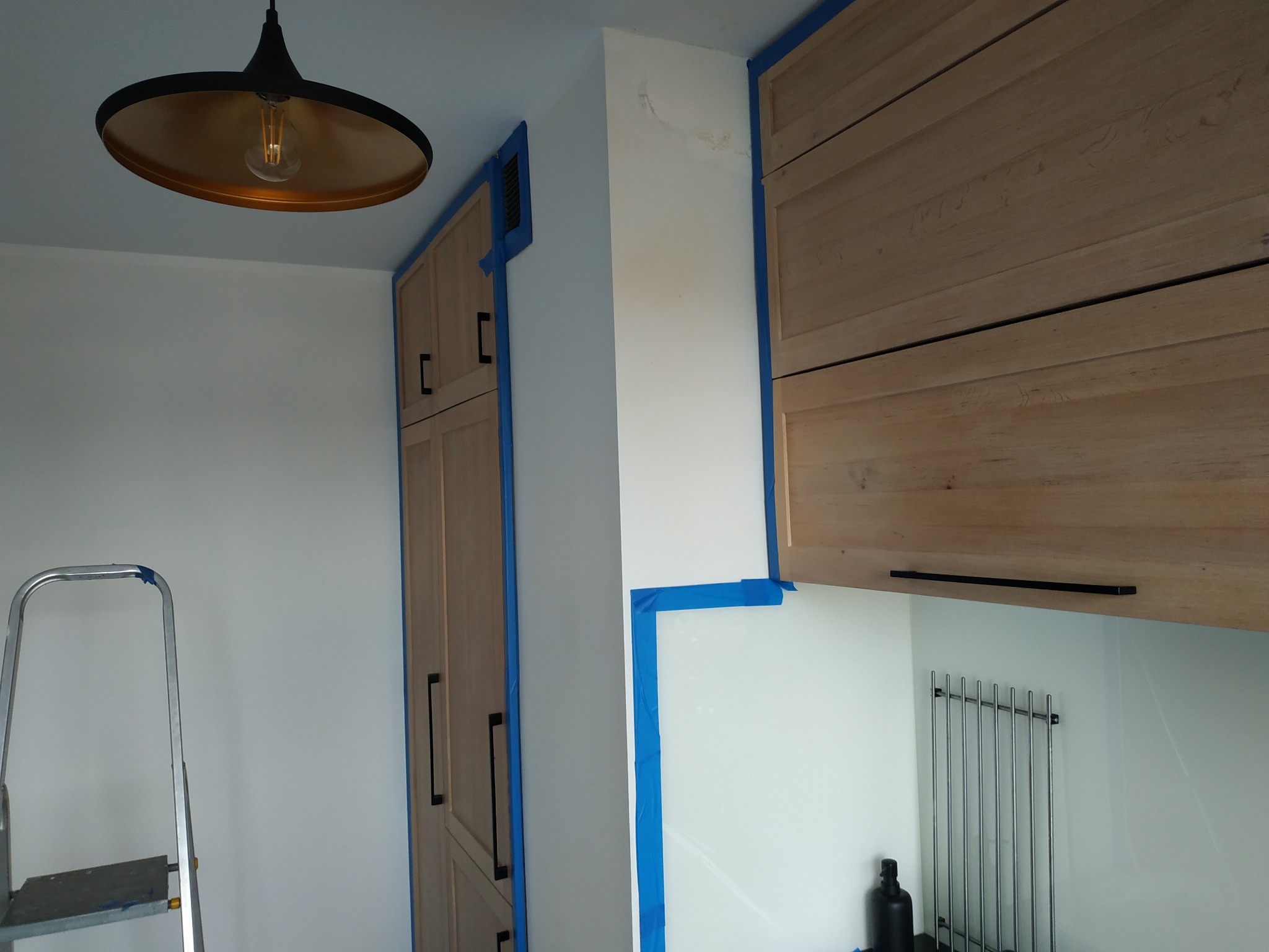 Przygotowania do prac wykończeniowych w kuchni: zabezpieczone taśmą meble i ściany, drabina oparta o ścianę, lampa wisząca z żarówką Edisona