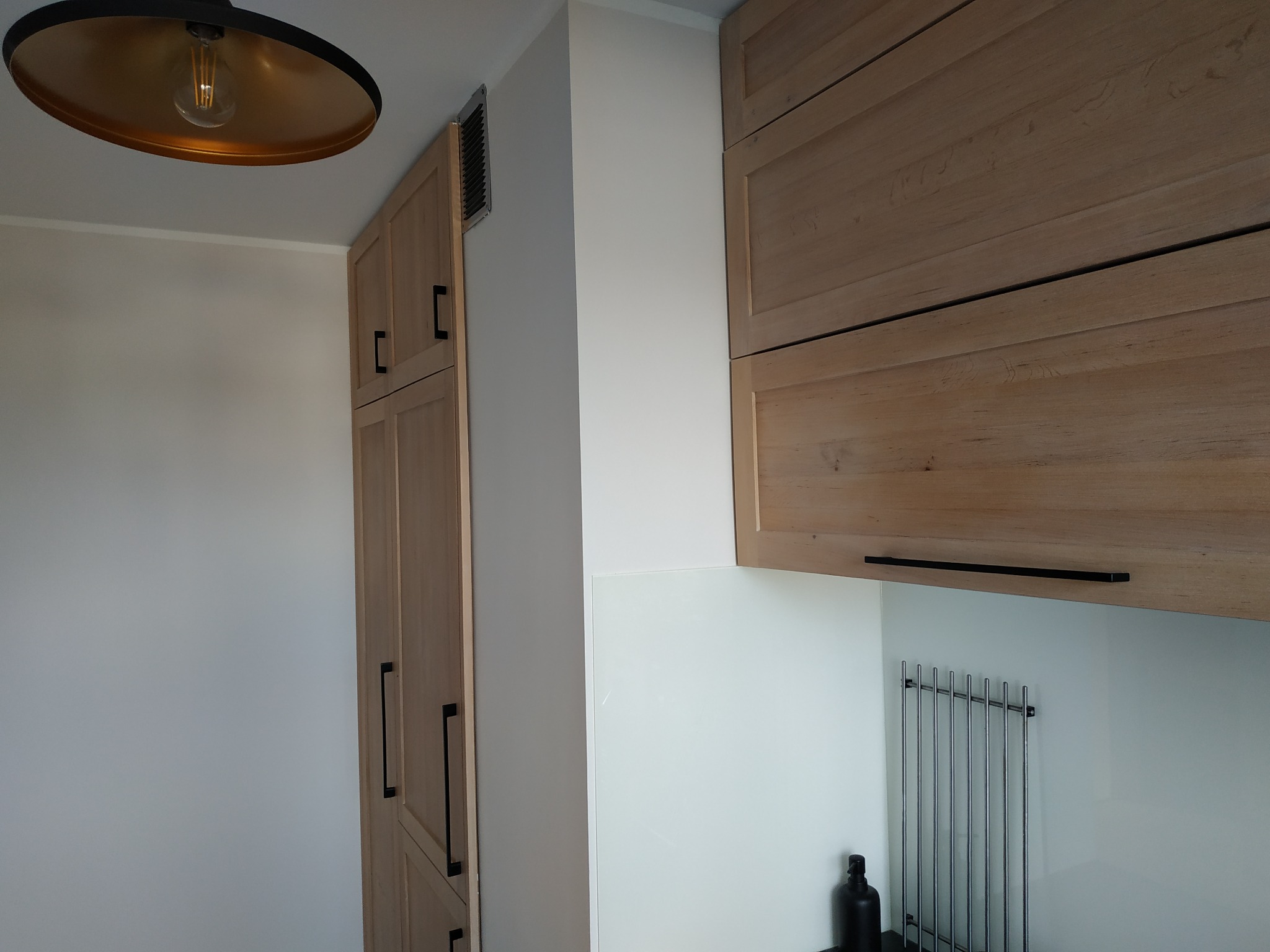 Jasne, drewniane szafki kuchenne z czarnymi uchwytami, białe ściany, lampa sufitowa z elementami miedzianymi i metalowa suszarka do naczyń, widok z dołu.