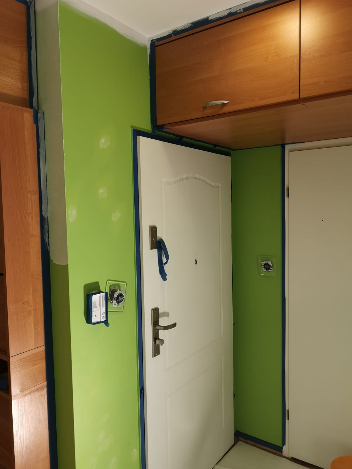 Ściana w trakcie odświeżania koloru na jaskrawy zielony, zabezpieczona taśmą malarską przy futrynach i szafkach, widoczne niedomalowane fragmenty i gniazdko elektryczne zdemontowane do prac.