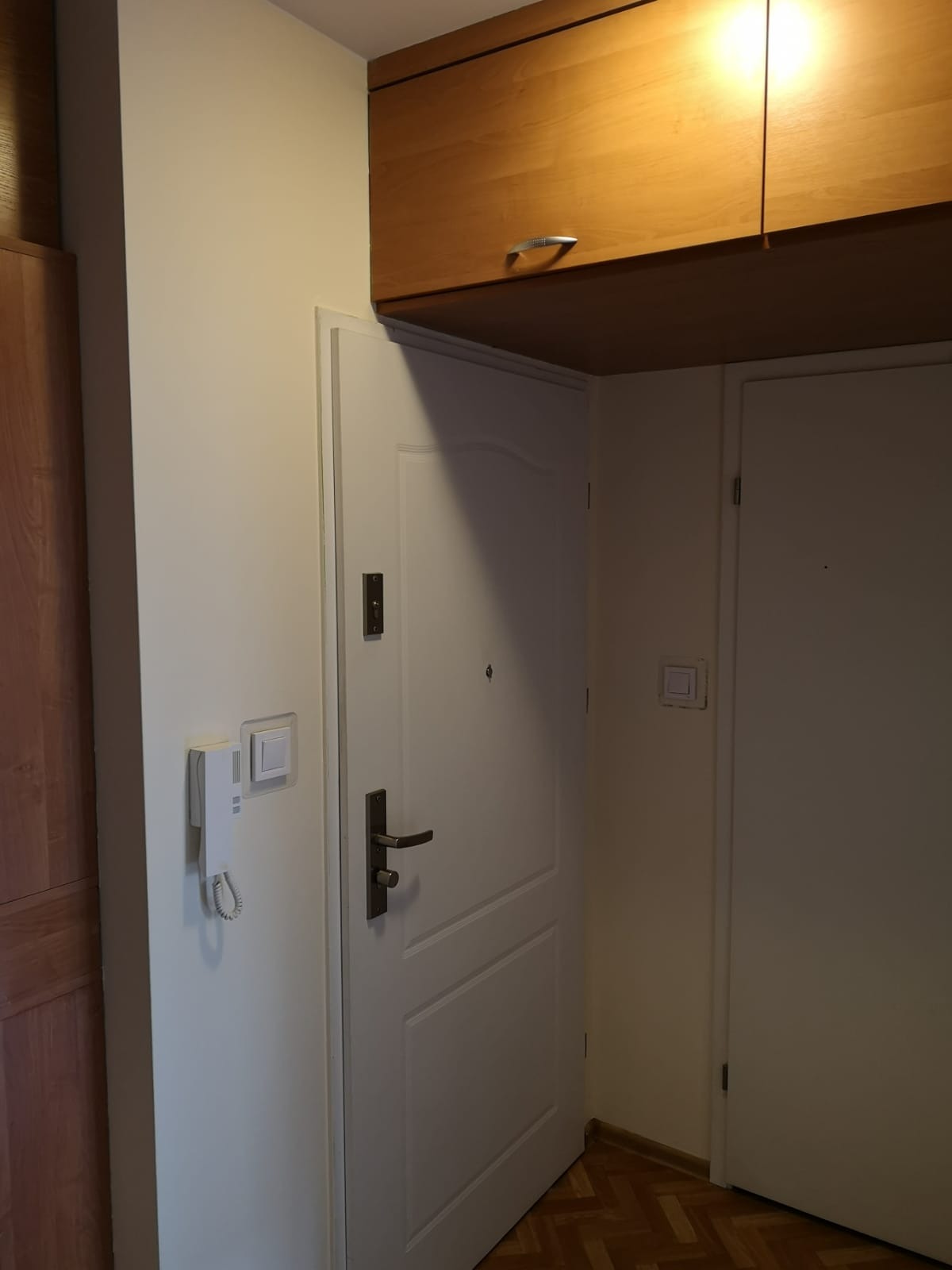 Białe ściany w przedpokoju po remoncie, widoczne drzwi wejściowe i szafka pod sufitem.