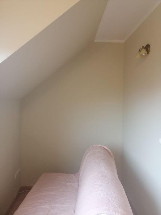 Pomalowane na beżowo poddasze ze skosem, widoczny fragment sofy w kolorze pudrowego różu i kinkiet z dwoma kloszami.