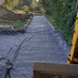 Przygotowanie terenu pod drogę: wykop wyłożony czarną folią, widoczna krawędź łyżki koparki, w tle drzewa i pryzma ziemi.