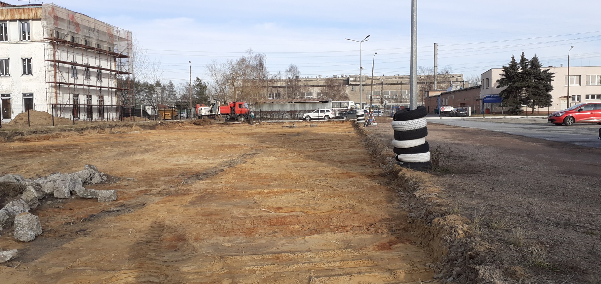 Przygotowany teren pod budowę z widocznym wykopem, stosem opon przy krawędzi i pracującą koparką w tle, obok budynku w rusztowaniach.
