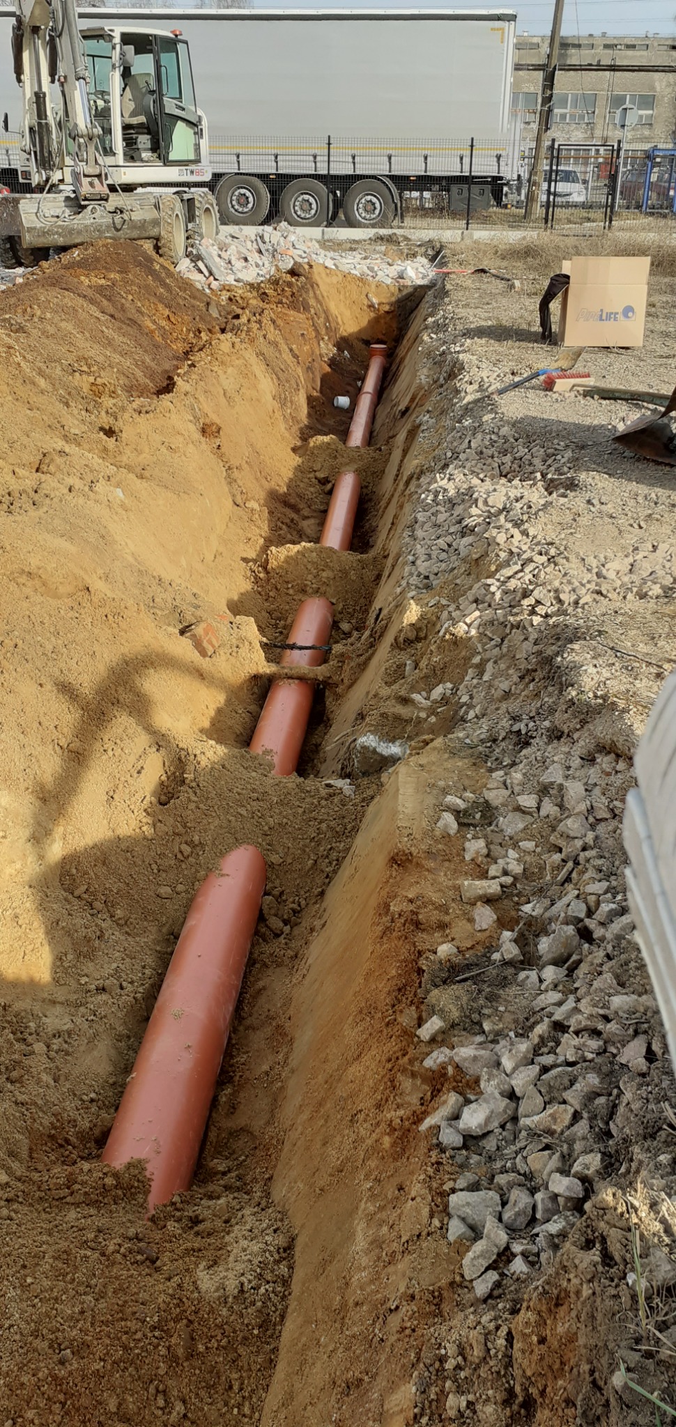 Głęboki wykop w ziemi z ułożonymi rurami kanalizacyjnymi z tworzywa w kolorze ceglastym; na górze widoczny fragment koparki i ciężarówki, a obok wykopu gruz i kamienie.