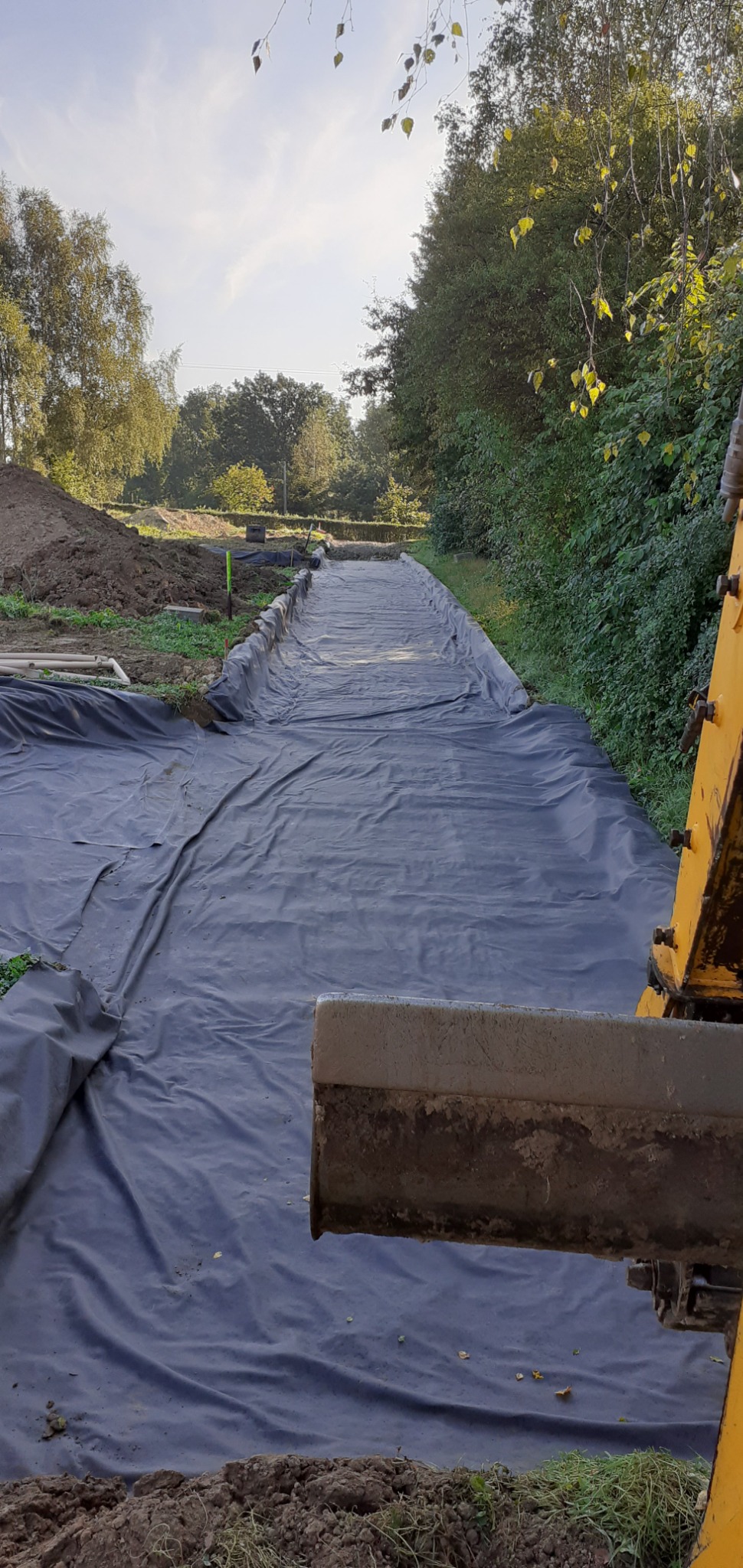 Przygotowanie terenu pod drogę: wykop wyłożony czarną folią, widoczna krawędź łyżki koparki, w tle drzewa i pryzma ziemi.