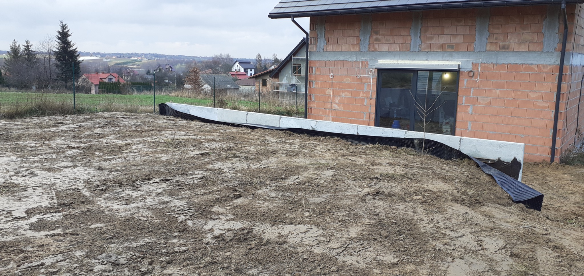 Fundament budynku w stanie surowym otoczony izolacją przeciwwilgociową i drenażem, widok na posesję i okoliczne zabudowania w tle.