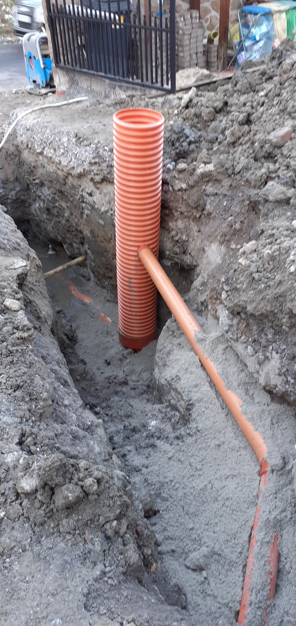 Pomarańczowe rury karbowane ułożone w wykopie ziemnym, widoczny fragment instalacji podziemnej z podłączeniem rury poziomej, na drugim planie ubijarka i ogrodzenie.