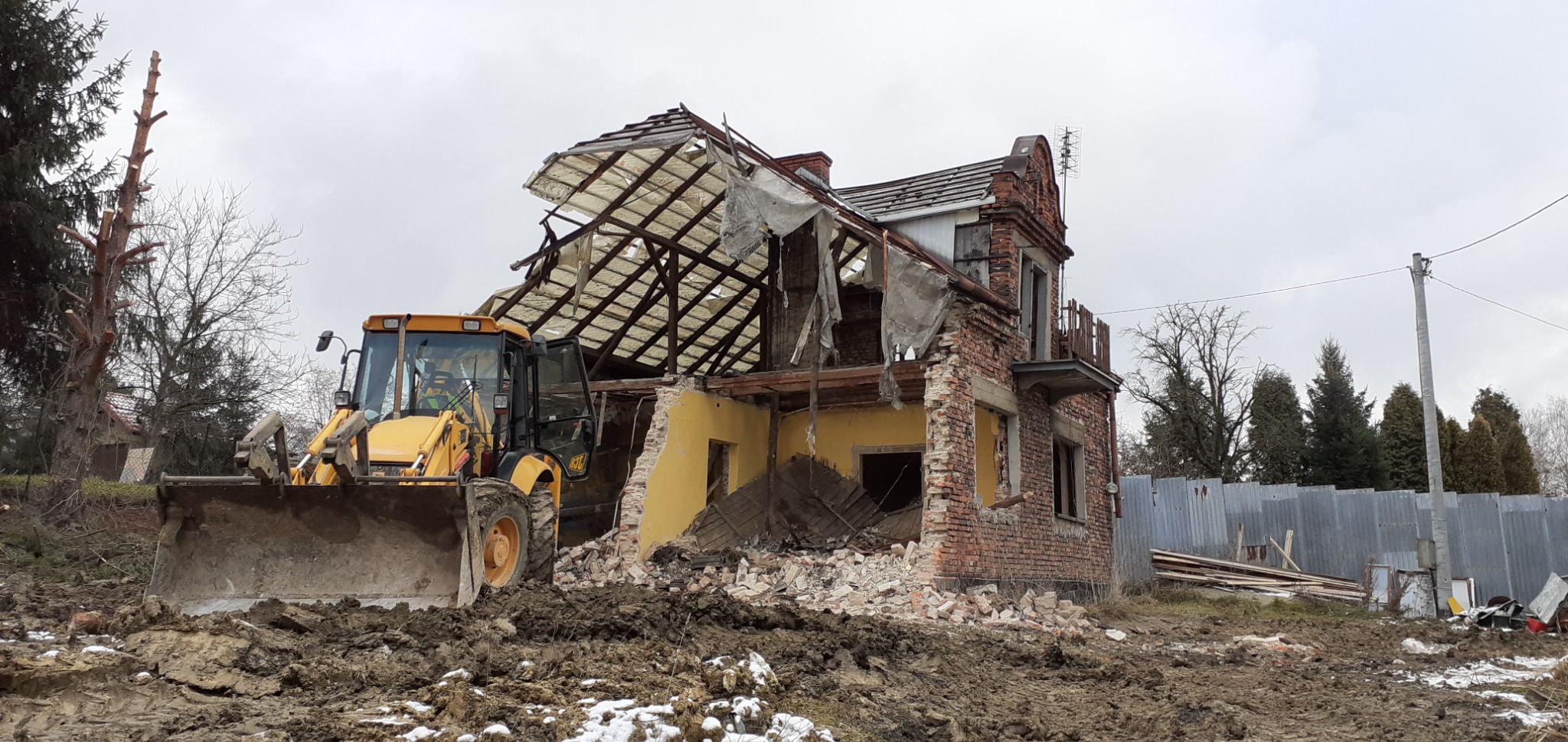 Żółta koparko-ładowarka na błotnistym terenie przed częściowo zburzonym, dwupiętrowym budynkiem z cegły w Krakowie. Widoczne odsłonięte elementy konstrukcyjne dachu i fragmenty ścian.