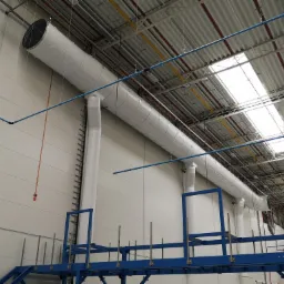 Biały, segmentowy kanał wentylacyjny pod stropem hali przemysłowej, widoczny na tle białych ścian i metalowej konstrukcji pomostu w kolorze niebieskim.