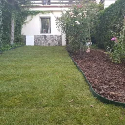 Świeżo założony trawnik z wyraźnie oddzieloną rabatą z kory, obrzeża z tworzywa sztucznego, w tle budynek z kamienną podmurówką i bujna roślinność.