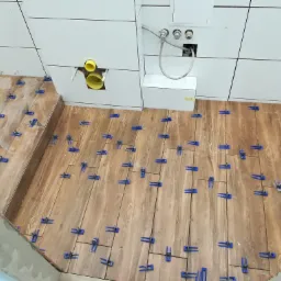 Proces układania płytek imitujących drewno na podłodze w Chorzowie, z użyciem niebieskich klipsów poziomujących, widoczne białe płytki na ścianie i instalacja sanitarna.