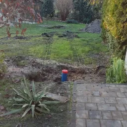 Ogród po wykopie, widoczna rura z niebieskiego plastiku z czerwoną nakrętką wystająca z ziemi, ślady opon i stos ziemi w tle, obok kostki brukowej i ściany budynku.