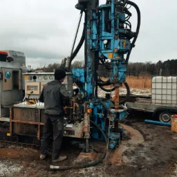 Operator w czapce obsługuje wiertnicę hydrauliczną w terenie z resztkami śniegu, widoczny przyczepiony zbiornik na wodę i błoto wokół wiertła.