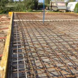 Zbrojenie płyty fundamentowej z drewnianym szalunkiem i podpory na piasku, w tle przyczepy kempingowe i zieleń.