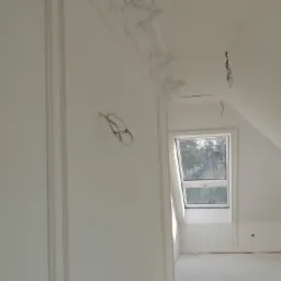 Wnętrze poddasza po remoncie, białe ściany z dekoracyjną sztukaterią, widoczne przewody elektryczne i okno dachowe.