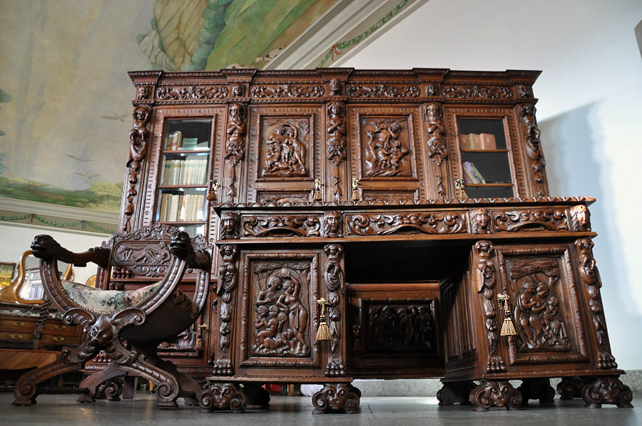 Bogato rzeźbiony, ciemnobrązowy komplet mebli gabinetowych w stylu neorenesansowym, składający się z biurka z nadstawką biblioteczną i krzesła z lwimi łapami, na tle jasnej ściany z malowidłem.