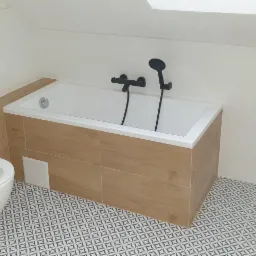 Nowoczesna łazienka na poddaszu z wanną obudowaną płytkami imitującymi drewno, wiszącą toaletą, czarną armaturą i pralką Samsung, wykończona geometryczną mozaiką podłogową.