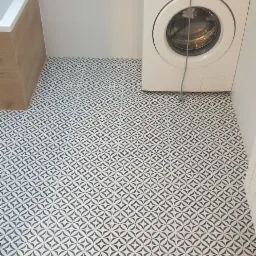 Ukończona podłoga w łazience z biało-czarną geometryczną mozaiką, widoczna pralka Samsung i fragment zabudowy wanny z drewnopodobną okleiną.