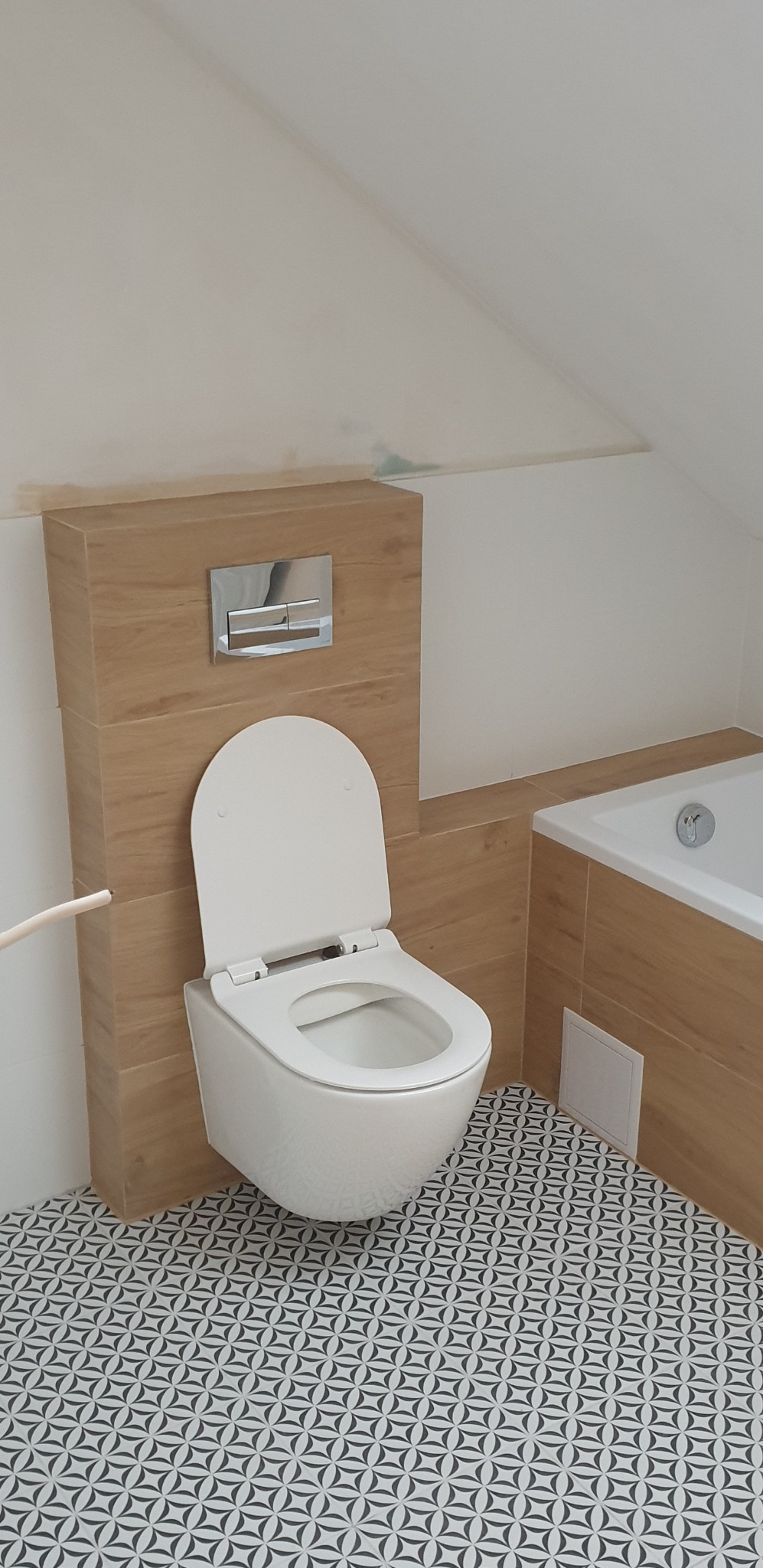 Nowoczesna, podwieszana toaleta z otwartą deską, zamontowana na tle drewnopodobnej zabudowy z chromowanym przyciskiem spłukującym, w łazience z biało-czarną wzorzystą podłogą i fragmentem wanny...