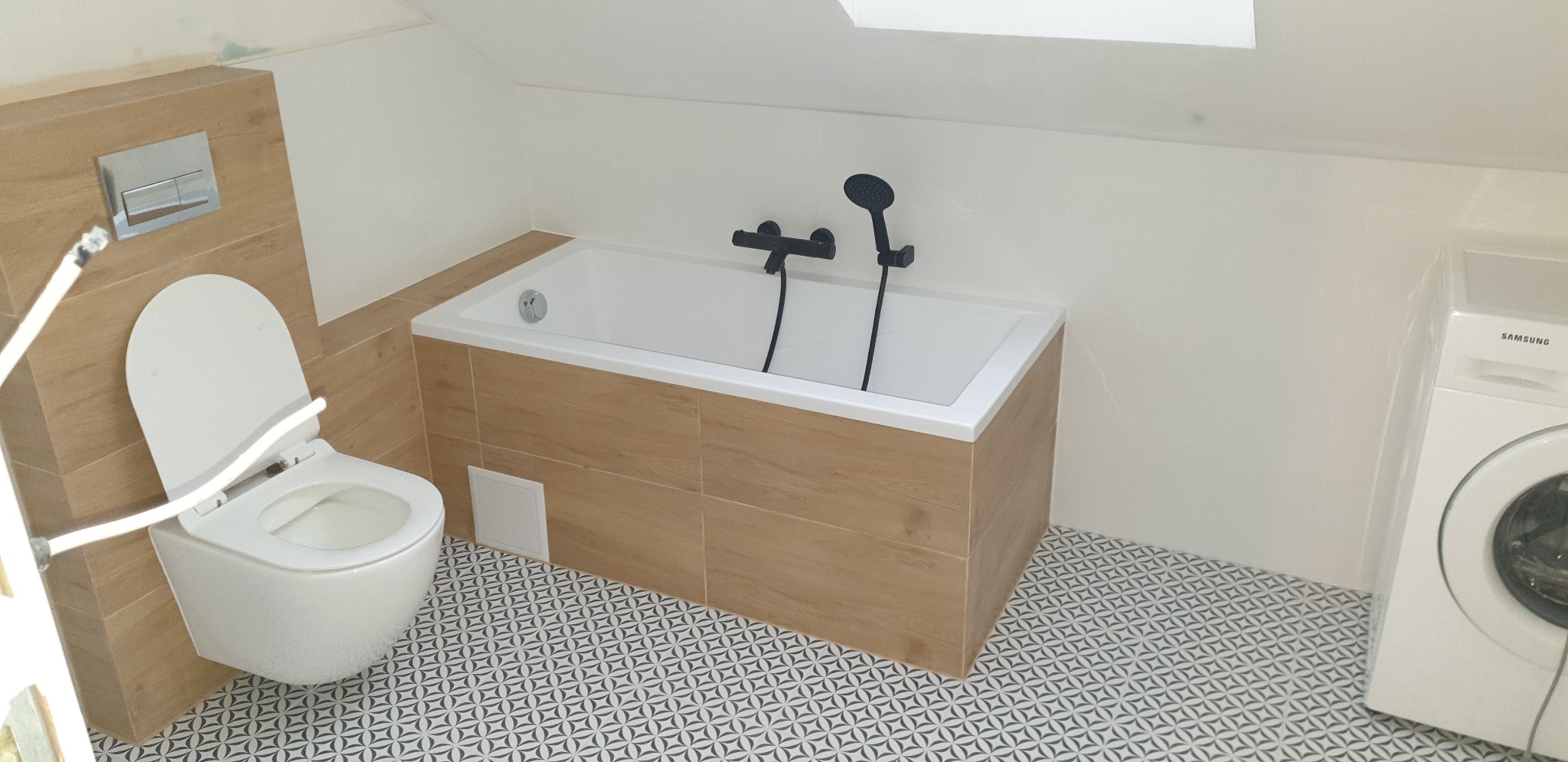 Nowoczesna łazienka na poddaszu z wanną obudowaną płytkami imitującymi drewno, wiszącą toaletą, czarną armaturą i pralką Samsung, wykończona geometryczną mozaiką podłogową.