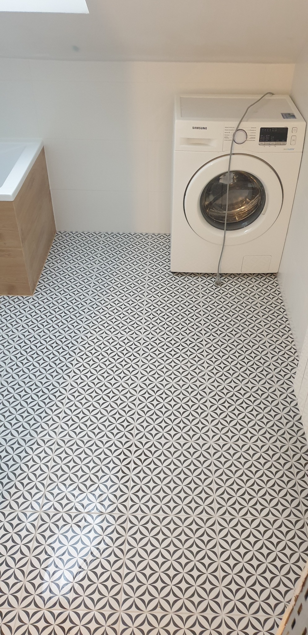 Ukończona podłoga w łazience z biało-czarną geometryczną mozaiką, widoczna pralka Samsung i fragment zabudowy wanny z drewnopodobną okleiną.