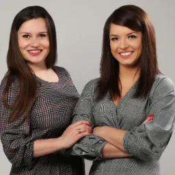 Portret dwóch uśmiechniętych kobiet w sukienkach w kratę, stojących na neutralnym tle. Jedna z założonymi rękami, druga z dłońmi splecionymi na jej ramieniu. Widoczny pierścionek na palcu jednej...