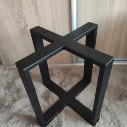 Czarna, metalowa podstawa stołu w kształcie litery X, wykonana w Rzeszowie, widoczna na tle puszystego dywanu i drewnianych drzwi przesuwnych.