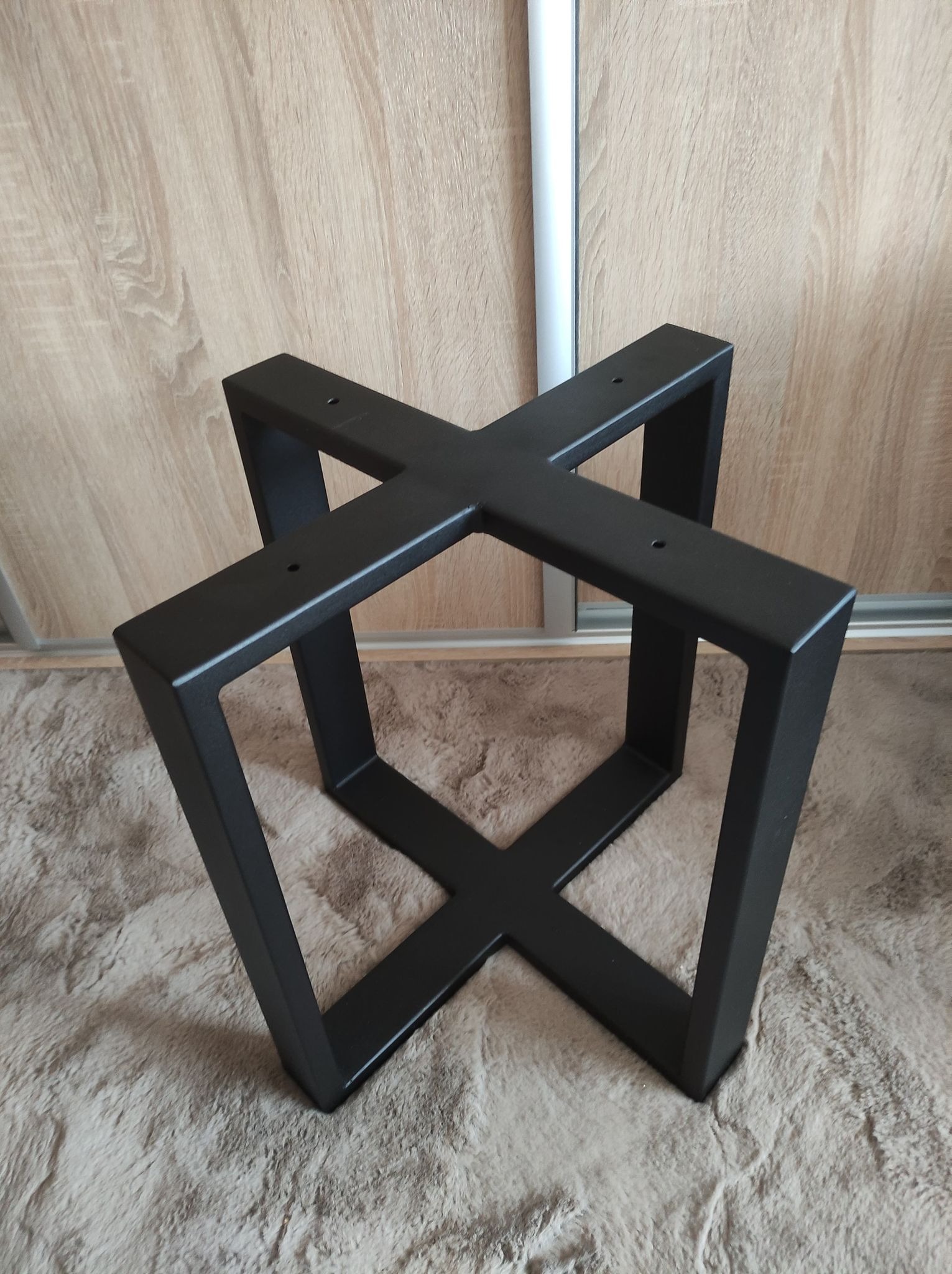 Czarna, metalowa podstawa stołu w kształcie litery X, wykonana w Rzeszowie, widoczna na tle puszystego dywanu i drewnianych drzwi przesuwnych.