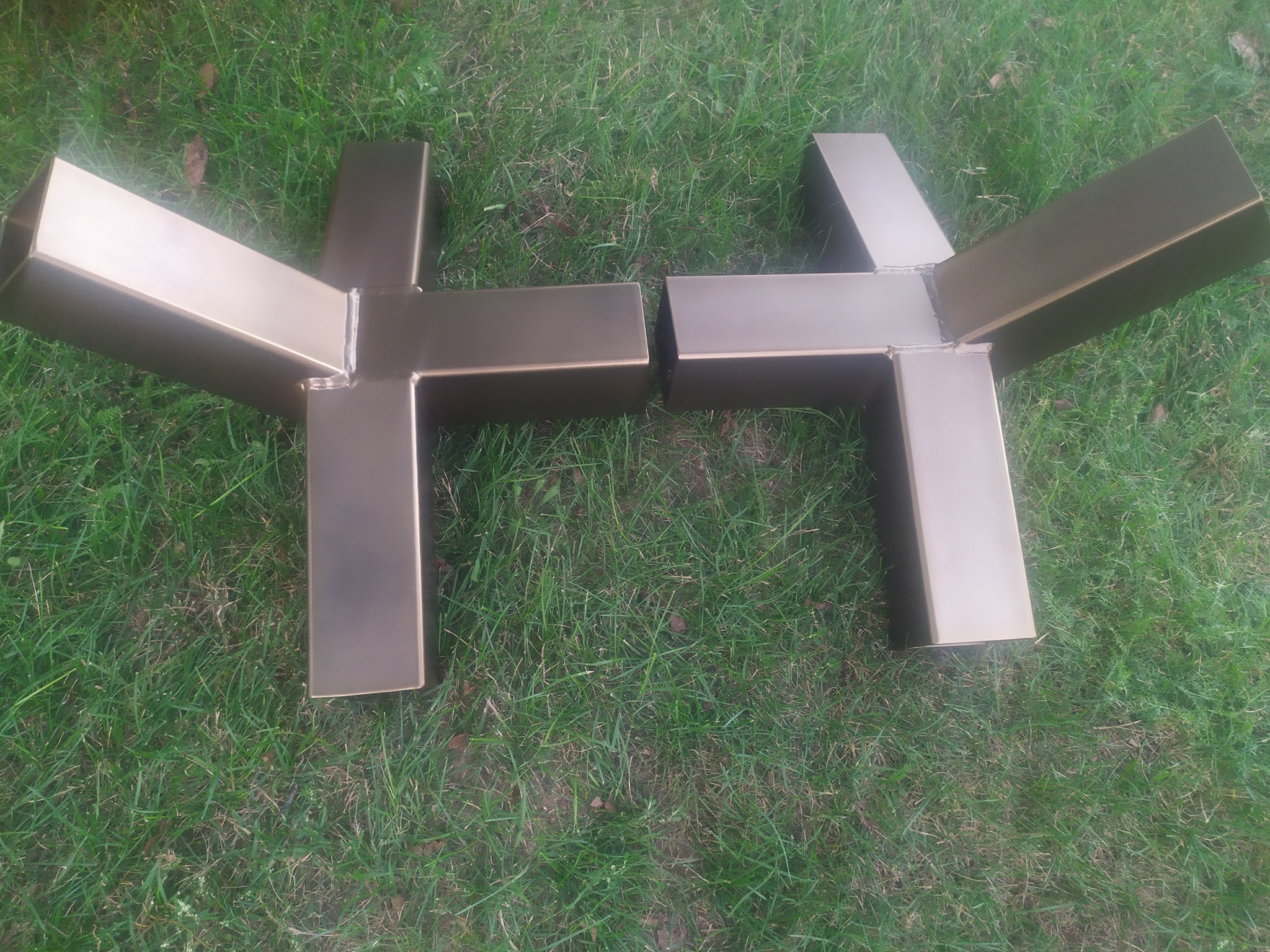 Dwa spawane, metalowe elementy konstrukcyjne w kształcie litery T, leżące na trawie. Widoczne spoiny na łączeniach.