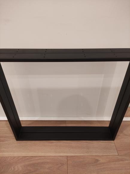 Czarna, spawana rama stołu z otworami montażowymi, ułożona na drewnianej podłodze na tle białej ściany.