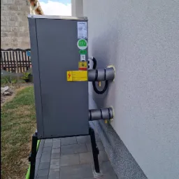 Szara jednostka zewnętrzna z podłączeniami do budynku, zamontowana na czarnych nóżkach, z widocznymi naklejkami informacyjnymi i zabezpieczeniami rur.