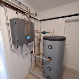 ML ENERGY SP. Z O.O. - Szary, wiszący piec z wyświetlaczem cyfrowym marki nesheat, połączony rurami z szarym, cylindrycznym zbiornikiem na wodę w pomieszczeniu z białymi kafelkami na ścianach.