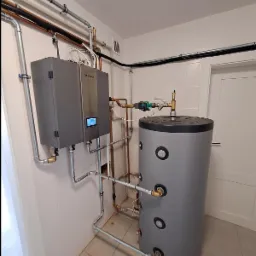 Szary, wiszący piec z wyświetlaczem cyfrowym marki nesheat, połączony rurami z szarym, cylindrycznym zbiornikiem na wodę w pomieszczeniu z białymi kafelkami na ścianach.