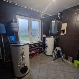ML ENERGY SP. Z O.O. - Kotłownia z dwoma zbiornikami, rozdzielaczem ogrzewania podłogowego i wiszącym kotłem, laptop na zbiorniku, drabina aluminiowa, okno z widokiem na deszcz.