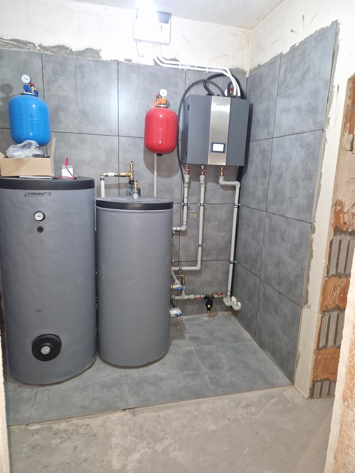 W kotłowni zainstalowano dwa szare zbiorniki akumulacyjne Trinnity, czerwony zbiornik przeponowy i szary moduł grzewczy z wyświetlaczem, połączone rurami z zaworami.