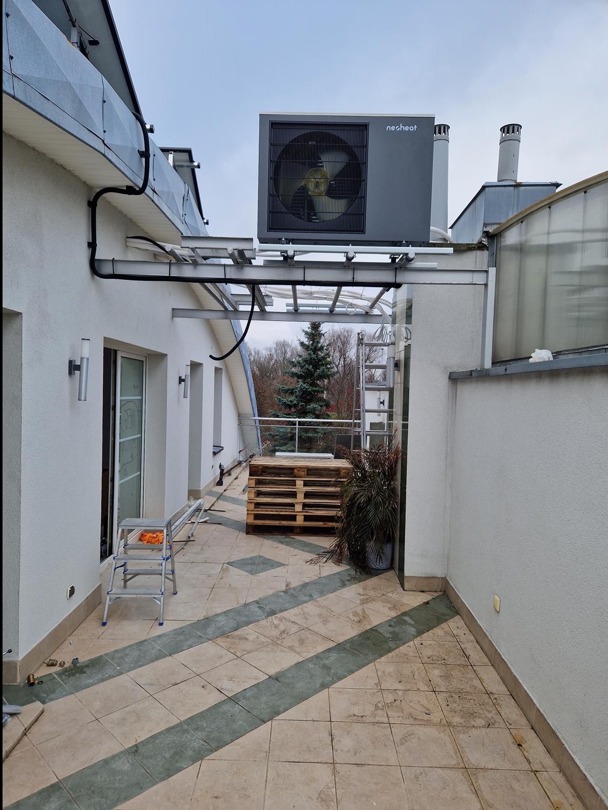 Szara jednostka zewnętrzna z wentylatorem, marki nesheat, zamontowana na metalowej konstrukcji na dachu budynku, widoczna drabina, palety i roślina w donicy na tarasie z płytkami w zielone pasy.