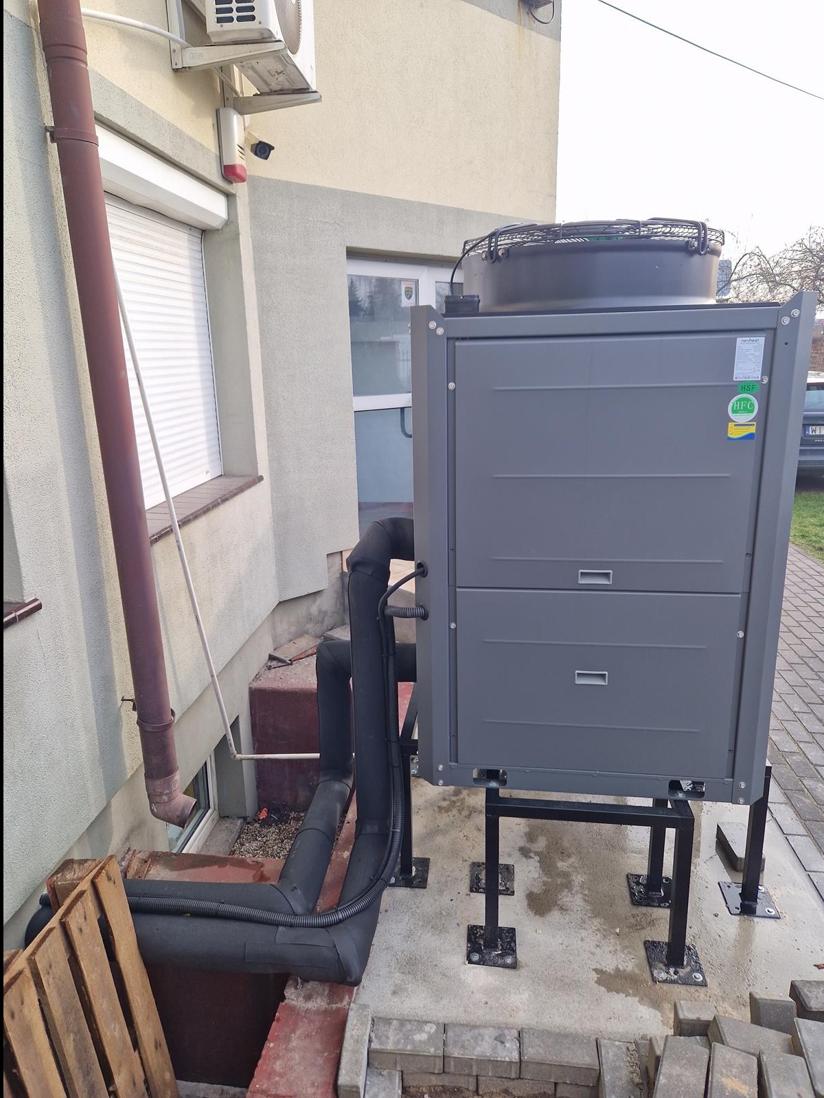 Szara pompa ciepła zamontowana na czarnych metalowych wspornikach obok budynku z widocznymi rurami izolacyjnymi i paletą drewnianą w tle.