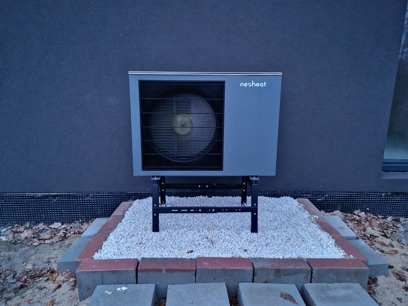 Szara jednostka zewnętrzna z widocznym wentylatorem i logo 'nesheat', zamontowana na czarnej metalowej konstrukcji, otoczona białym żwirem i obrzeżem z kostki, na tle ciemnoszarej ściany budynku.