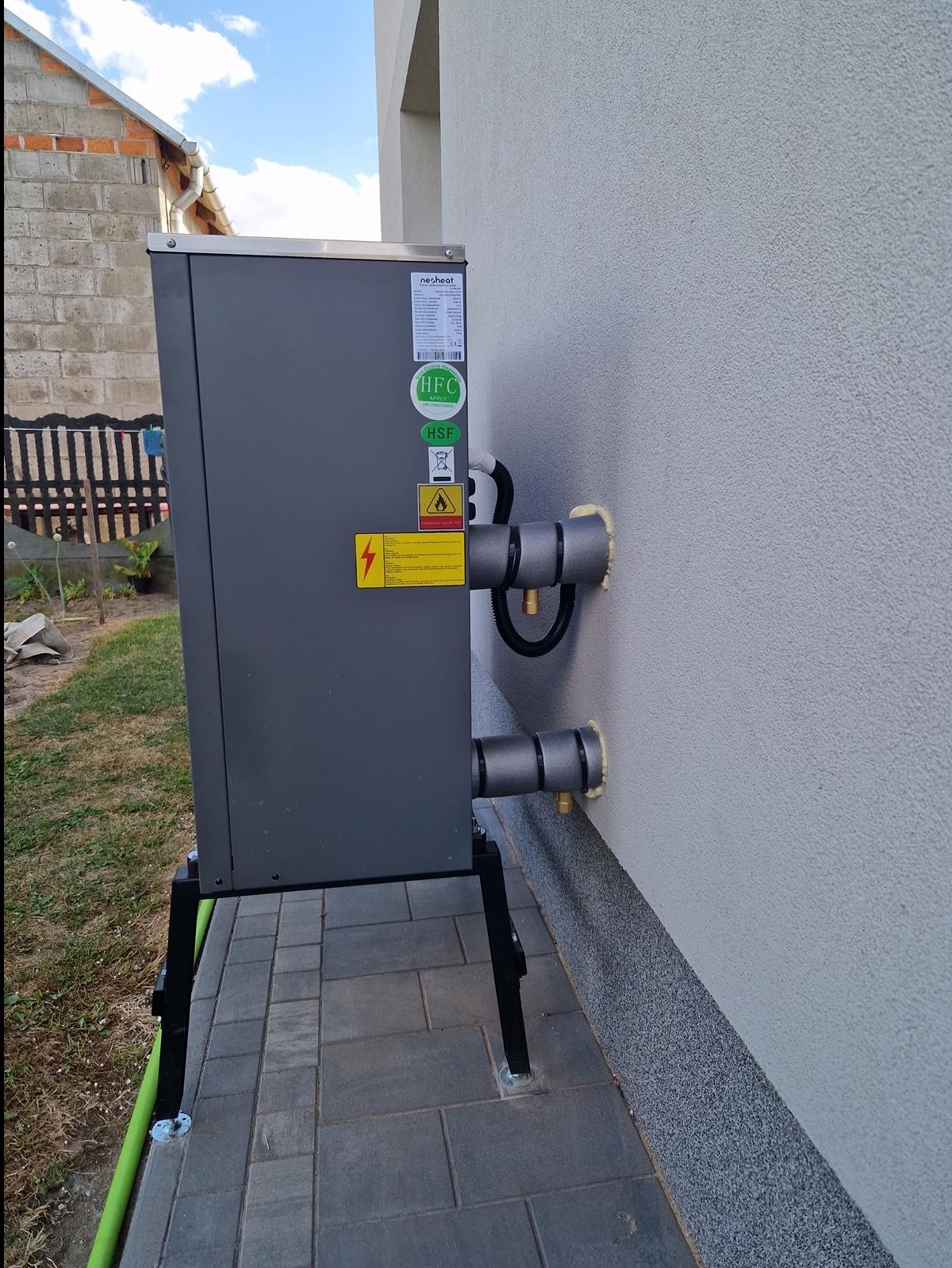 Szara jednostka zewnętrzna z podłączeniami do budynku, zamontowana na czarnych nóżkach, z widocznymi naklejkami informacyjnymi i zabezpieczeniami rur.