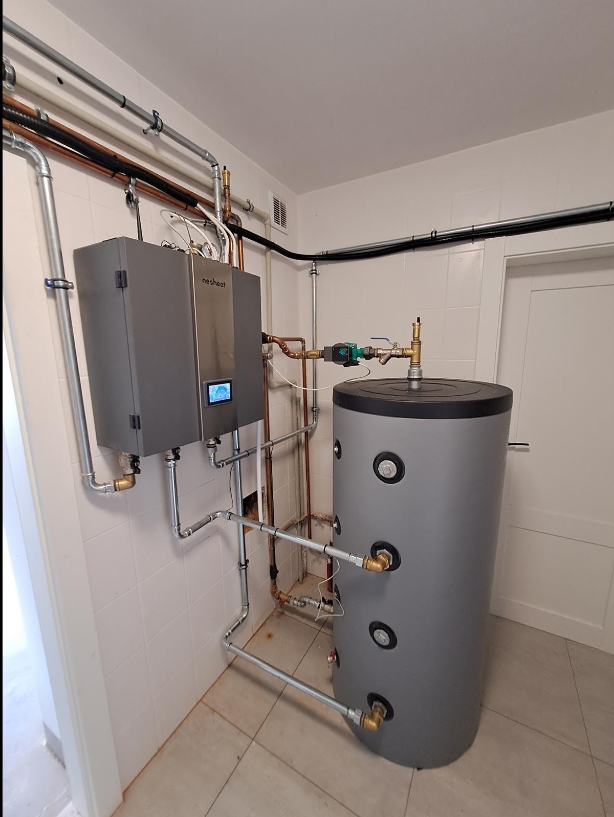 Szary, wiszący piec z wyświetlaczem cyfrowym marki nesheat, połączony rurami z szarym, cylindrycznym zbiornikiem na wodę w pomieszczeniu z białymi kafelkami na ścianach.