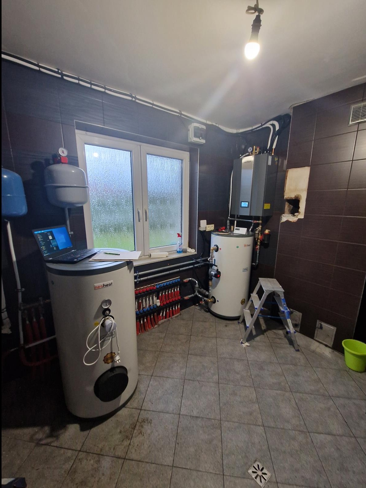 Kotłownia z dwoma zbiornikami, rozdzielaczem ogrzewania podłogowego i wiszącym kotłem, laptop na zbiorniku, drabina aluminiowa, okno z widokiem na deszcz.