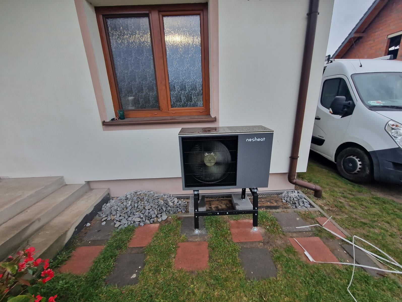 Zamontowana, szara pompa ciepła marki Nesheat na czarnym stelażu, widoczna obok ściany budynku z oknem, obok zaparkowany biały samochód dostawczy, na ziemi kamienie i trawa.